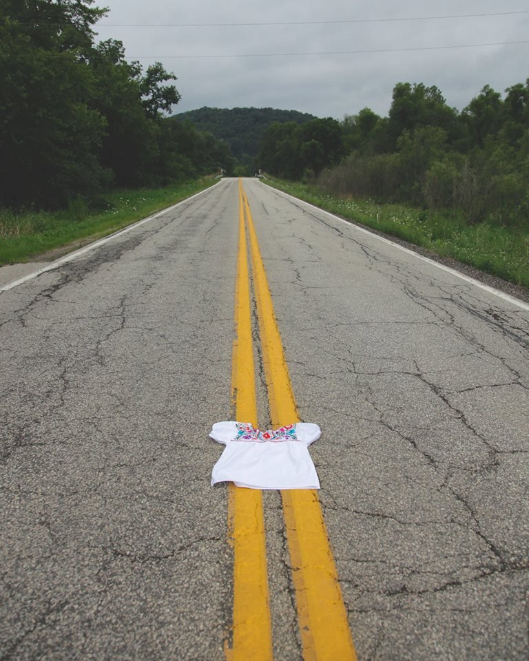 Double yellow lines stretch down a long road surrounded by dense green woods. In the foreground a white blouse with floral embroidery lays in the middle of the road. 