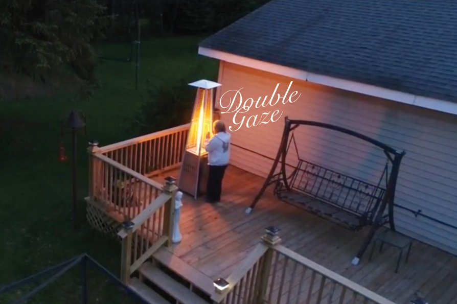 Screenshot seen from above showing a woman standing on a porch warming her hands on a propane furnace at dusk. 