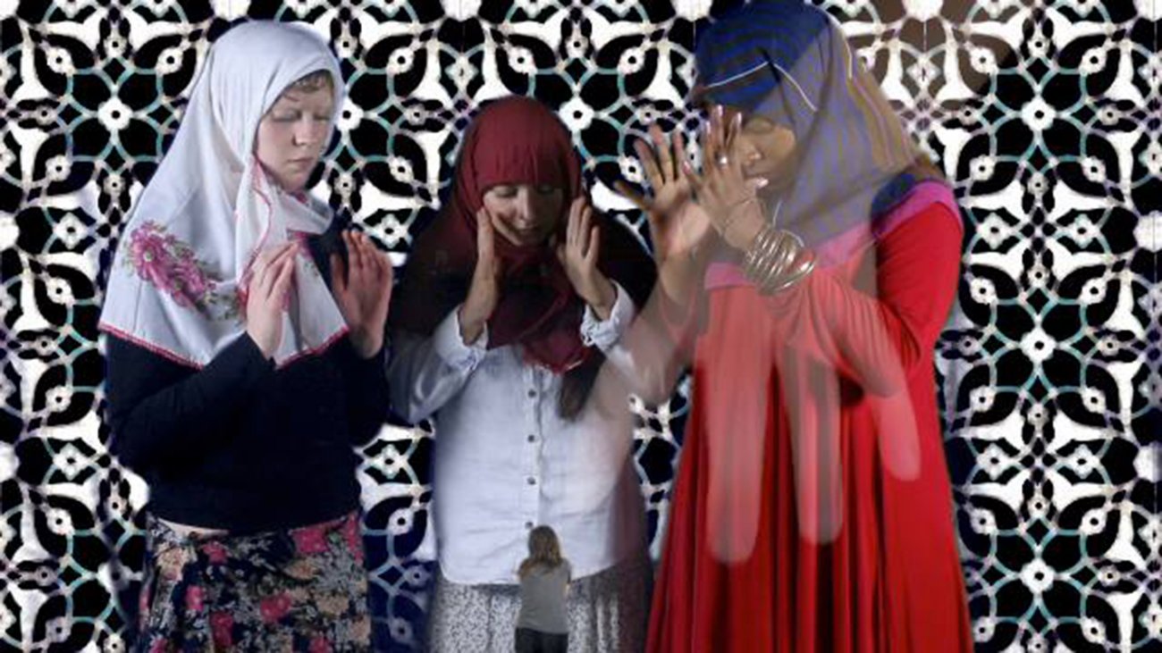 Three women wearing traditional head scarves stand with their hands in front of their chests as if they are about to carefully pick something up. They appear to be gathered around an animated image of the artist who is pictured as a the tiny subject of the women’s attention.