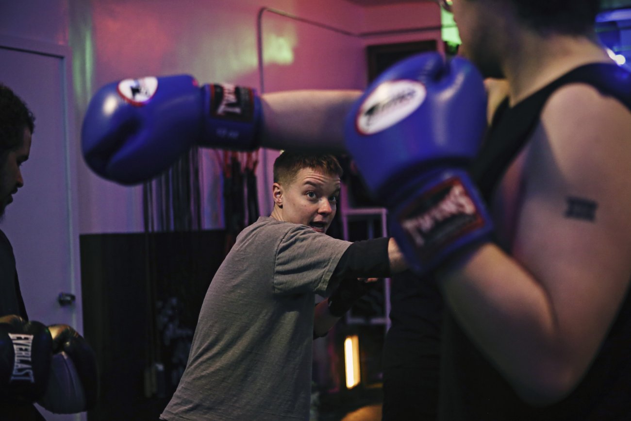 camera faces the artist who is in the center of the image standing in a boxing gym teaching a person on the right hand foreground of the image to throw a right cross.