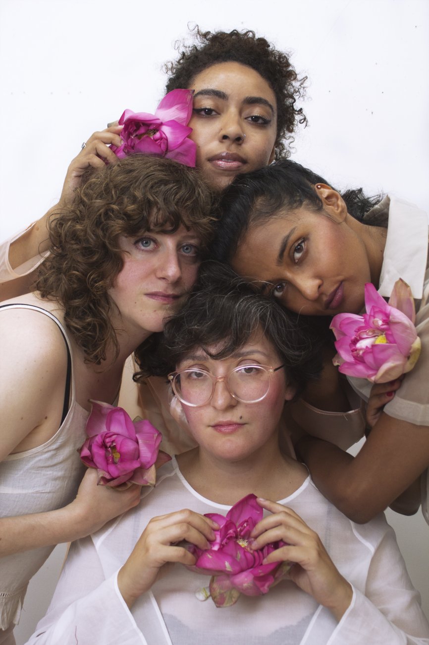 4 femme people dressed in white with their heads together in the center of the frame, each is holding a large pink flower
