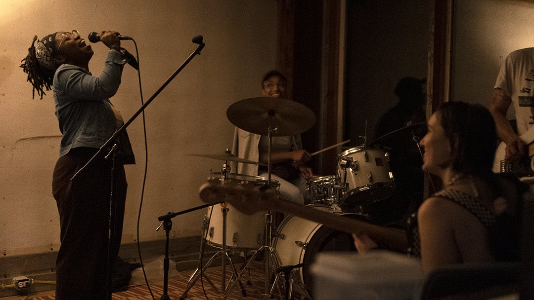 Femme presenting person with dark skin sings into a microphone in front of a band.