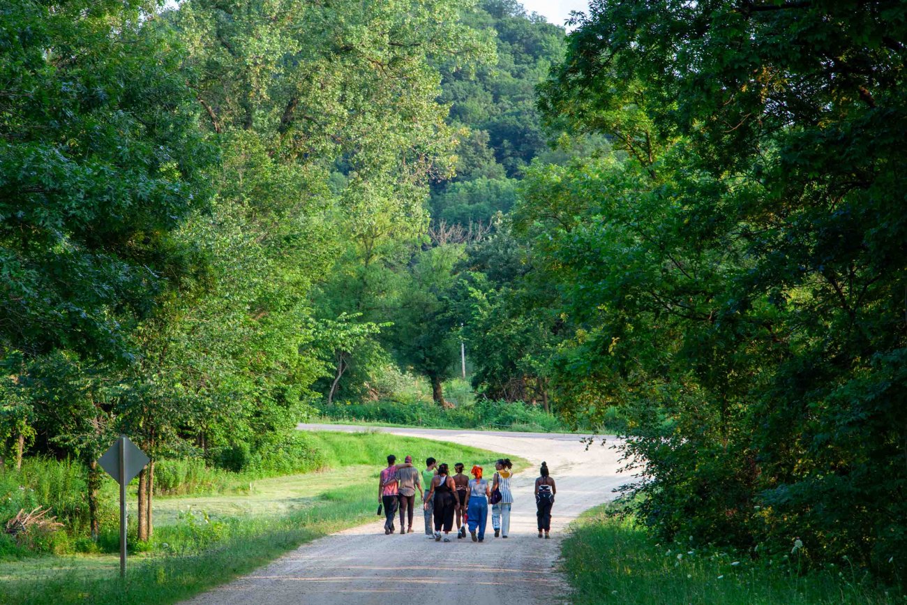 Group of artists walking down gravel road lined by vibrant green trees and grass (rear view)