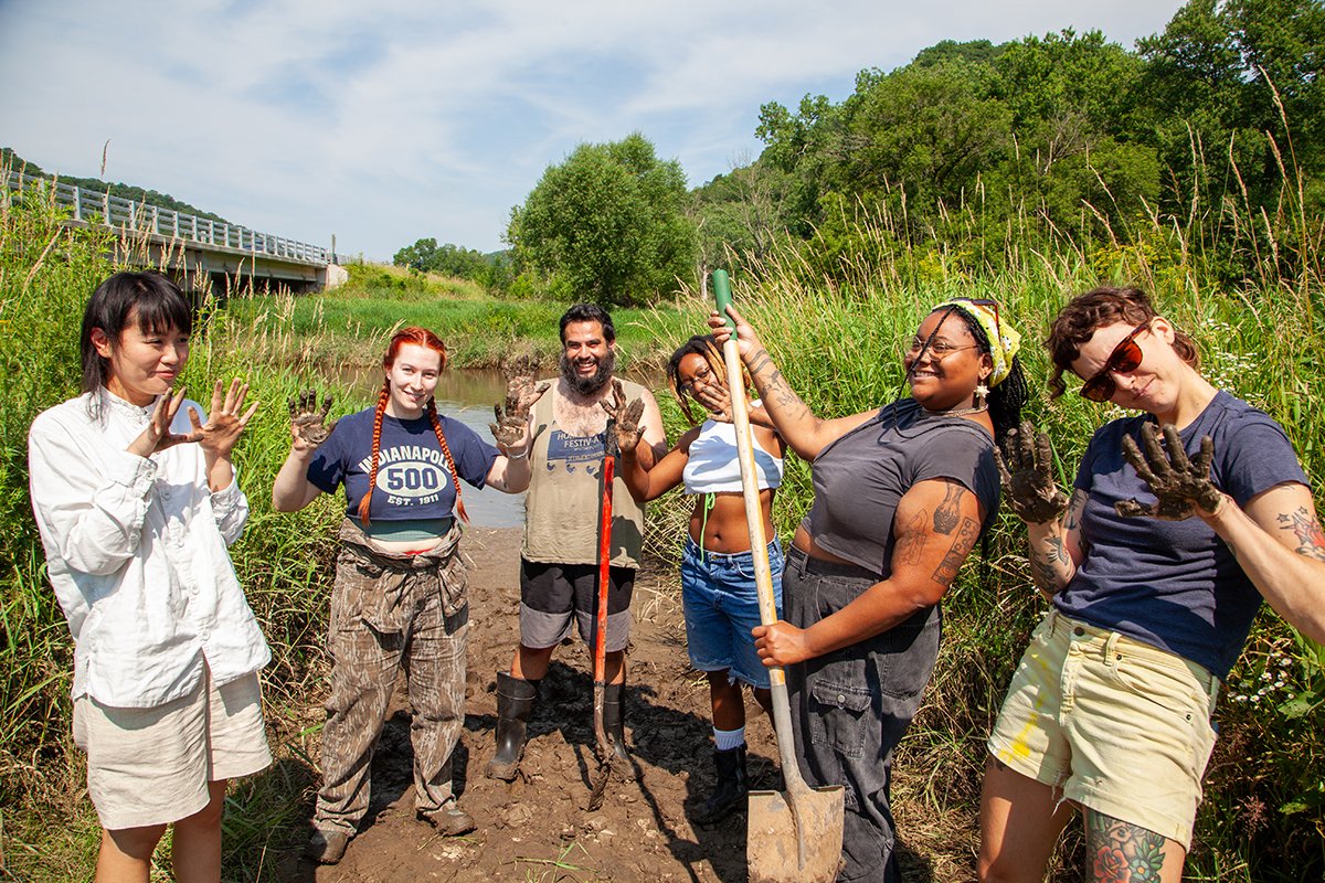 A group of artists show off their muddy hands as they dig by a riverbank for wild clay