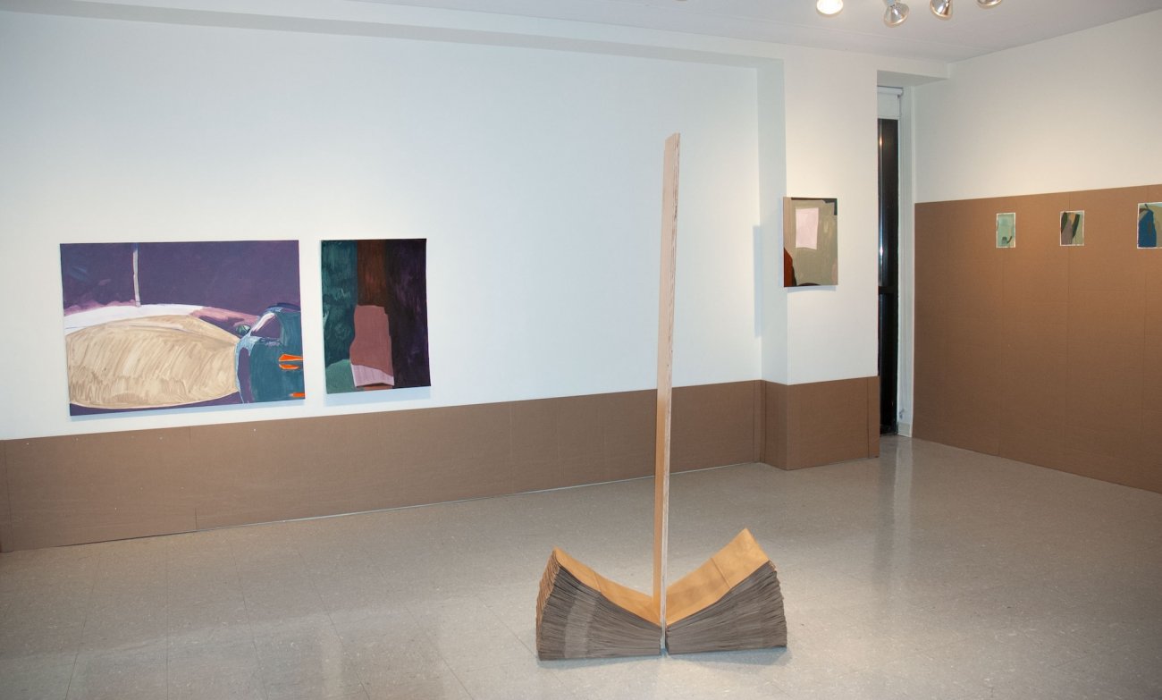 Two paintings are hung on the wall to the left with cardboard panels lining the wall just below. To the right of the same wall, another painting is displayed. On the adjoining wall, three small paintings are mounted on larger cardboard panels that cover about two-thirds of the wall. A three-dimensional piece, made up of two equal-sized stacks of paper bags is divided by an upright piece of wood, is in the foreground.