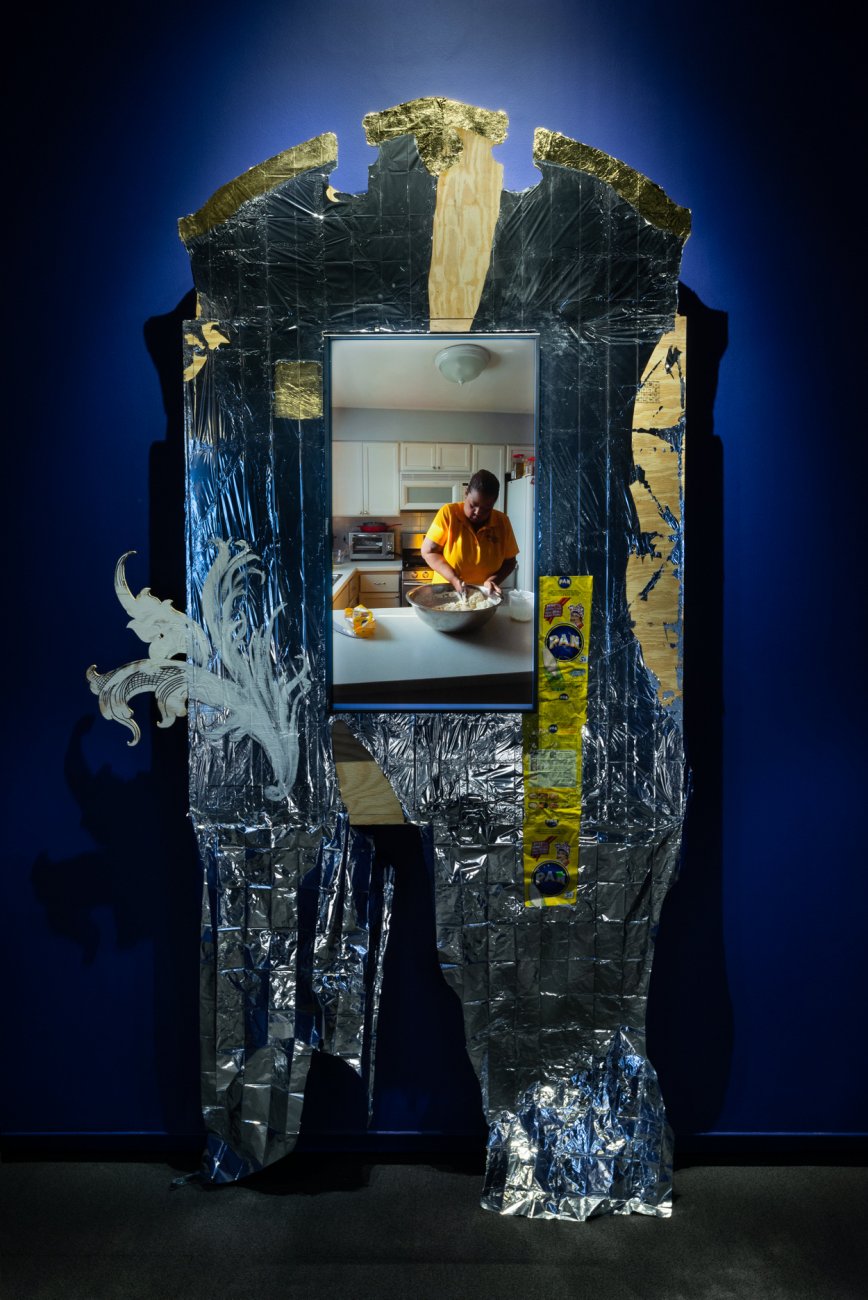 Darkly lit photograph of maylar blanket around a mirror reflecting a Black woman in a kitchen mixing flour in a large metal bowl