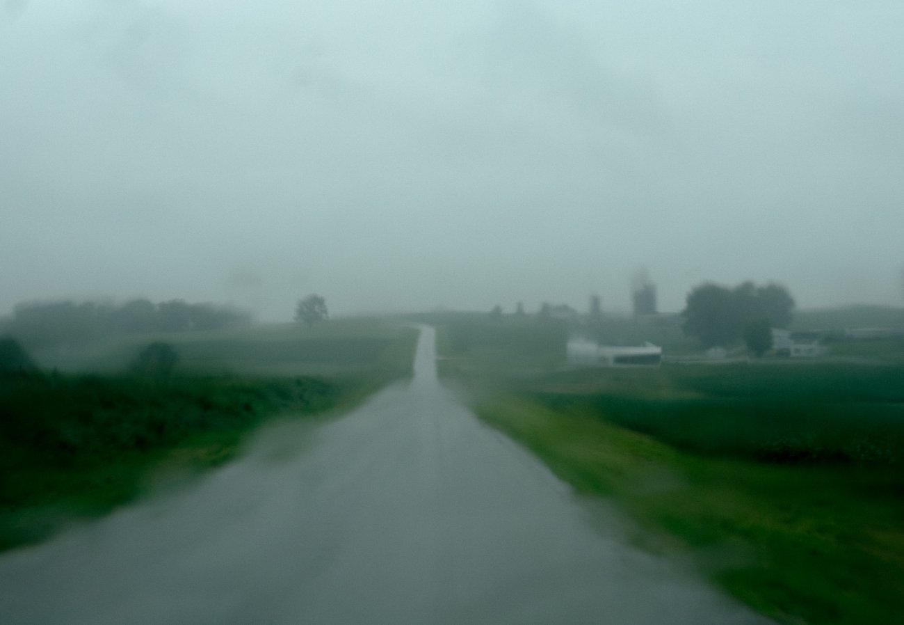 Fuzzy, gloomy photo of an empty wet road in the center of the frame with green fields on either side. Farm buildings and trees are in the distance.