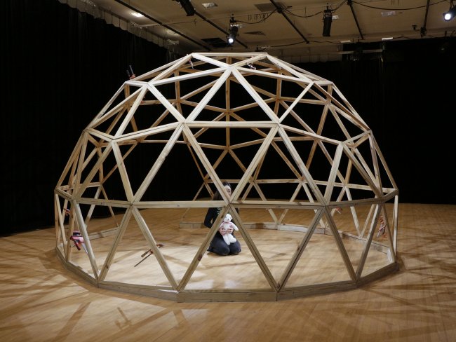 Photograph of a large geodesic dome made of wood, a person is crouched on their knees inside of the dome holding a stuffed animal