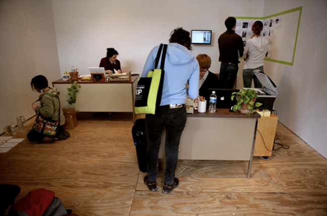 Documentation of in-person art reviews with two desks and various people around the room