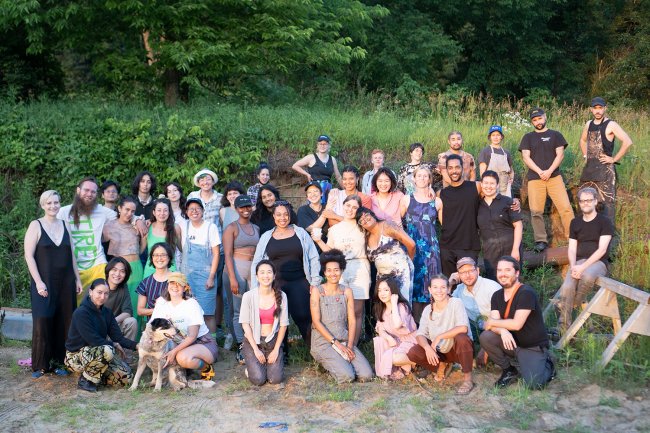 A group of smiling artists pose for an end-of-session portrait in front of tall grass and sloping trees