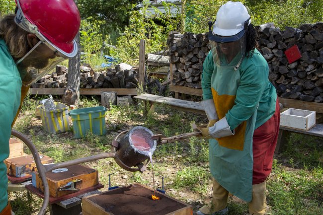 Two people in safety gear pouring liquid aluminum from a vessel into a sand cast mold