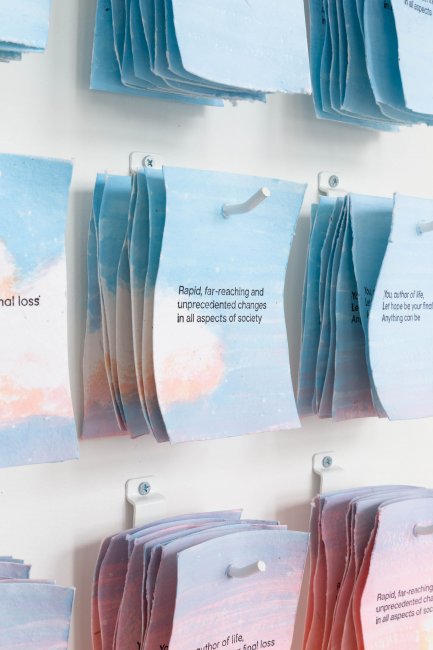 Installation view of artwork hanging on pegs. The center work reads "Rapid, far-reaching and unprecedented changes in all aspects of society"