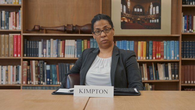 Still from "Committee of Six." A Black woman wearing glasses and a blazer stares directly at the camera. She sits at a desk in front of wall-to-wall bookshelves. A name plate reading "Kimpton" is on the table in front of her.