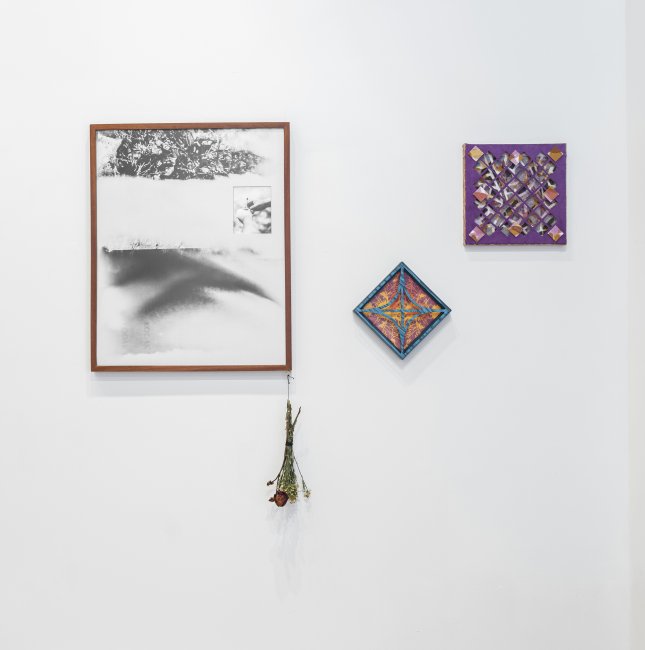 Black and white abstract image framed in a thin wood frame with a dead bouquet of flowers hanging off the bottom right corner. To the work's right are two small sculptural collages with paper cutouts.